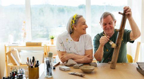 An older couple sits at a table by a window in their Life Plan Community, smiling as they work on a wooden craft project together. Paint and art supplies are spread out on the table, and sunlight fills the room.